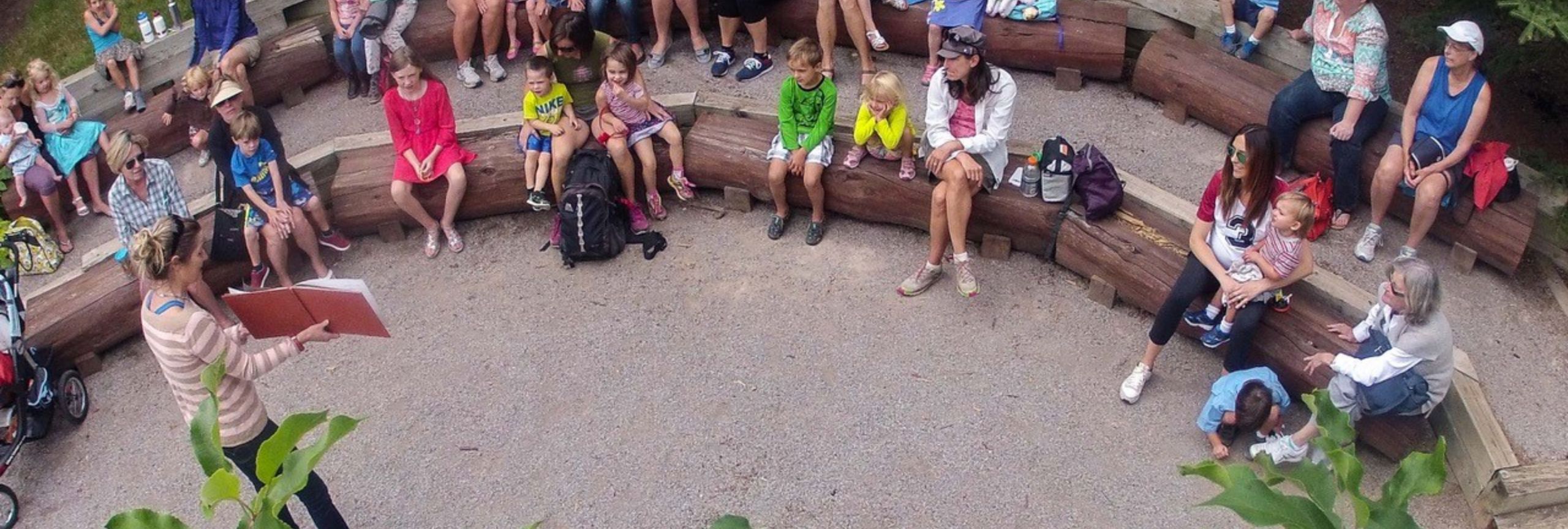 group of adults and children listening to woman reading from red book