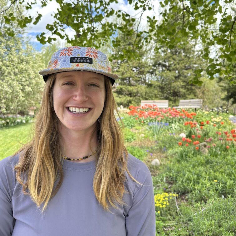 A blond woman with floral print hat standing in front of garden of wildflowers