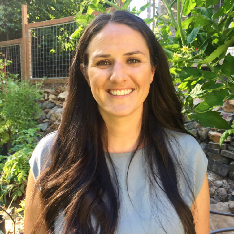 A woman with long dark hair smiles with the gate to Yampa River Botanic Park behind her