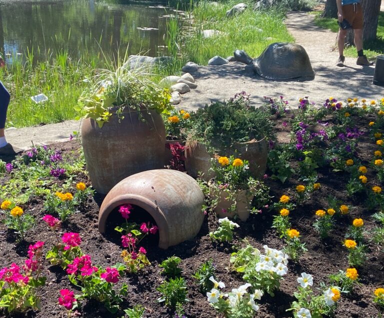 Rustic clay pots surrounded by blooming marigolds, petunias, and other annuals in a well-tended flower bed by a pond.