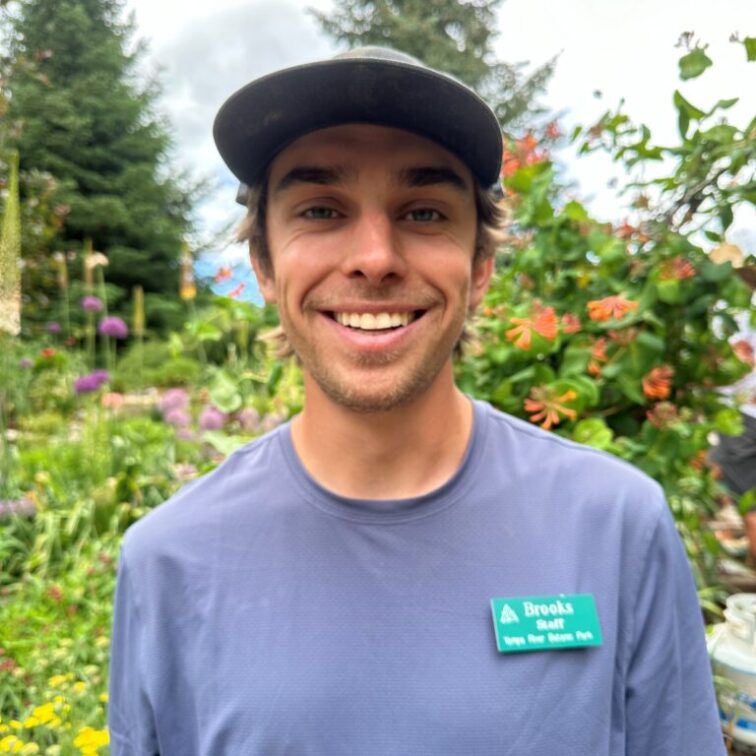 A dark haired man with floral print hat standing in front of garden of wildflowers