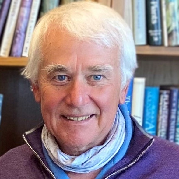 A man with white hair smiles against a background with full bookshelves