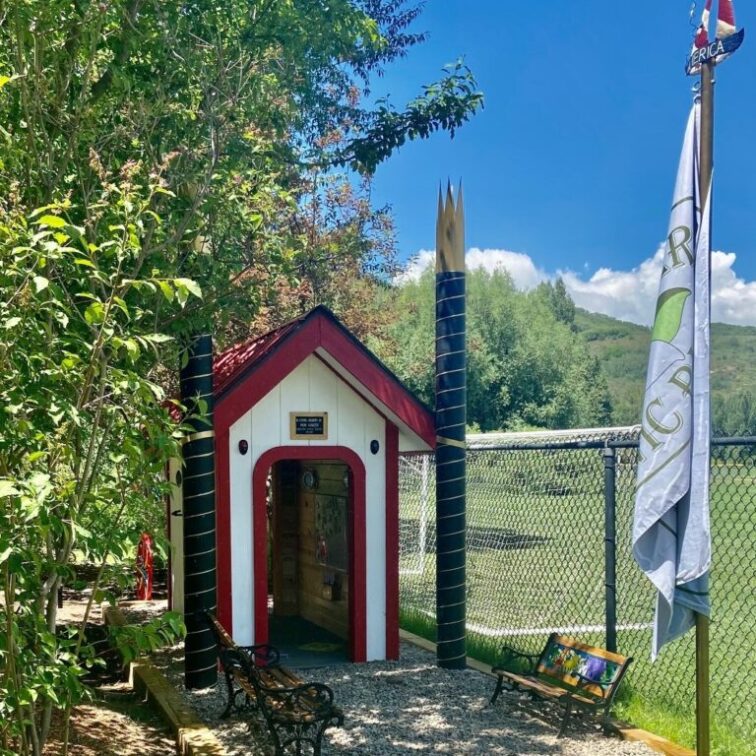 A small white and red structure with child sized benches near then entrance and large trees providing shade
