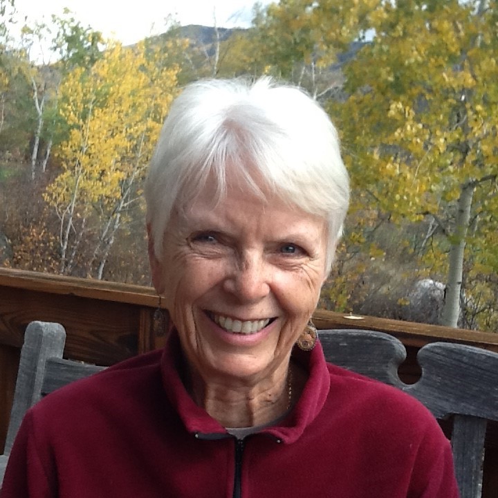 A woman with silver hair and a red sweater sits happily on a bench with yellow aspen trees in the background