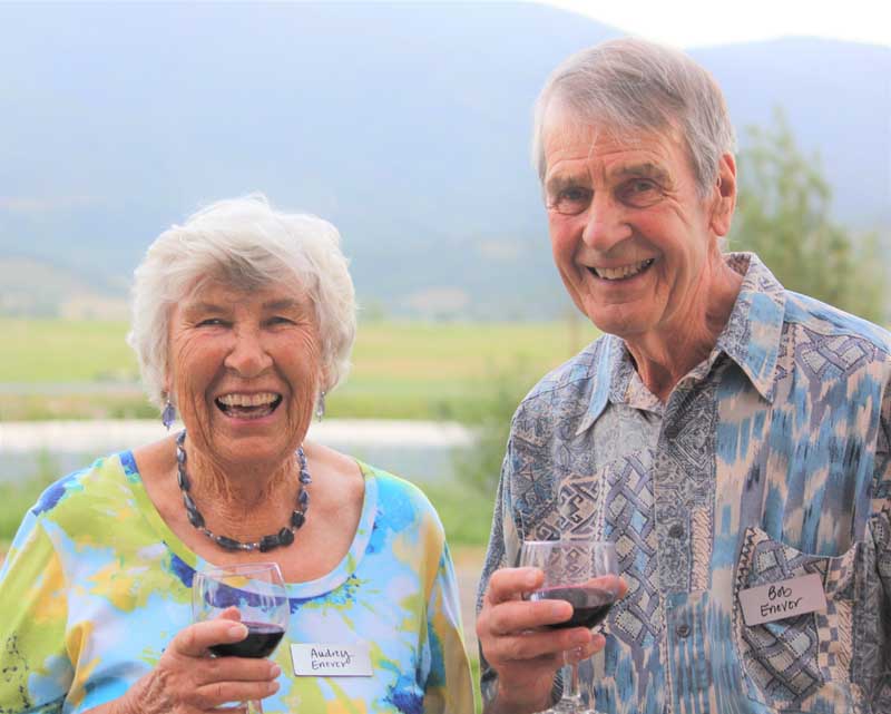 Bob and Audrey Enever stand together toasting with glasses of wine with mountains in the background