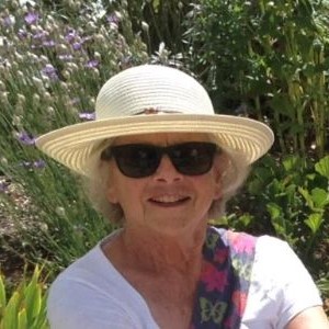 A blond woman wearing sunglasses and a sunhat smiles with purple flowers in the background