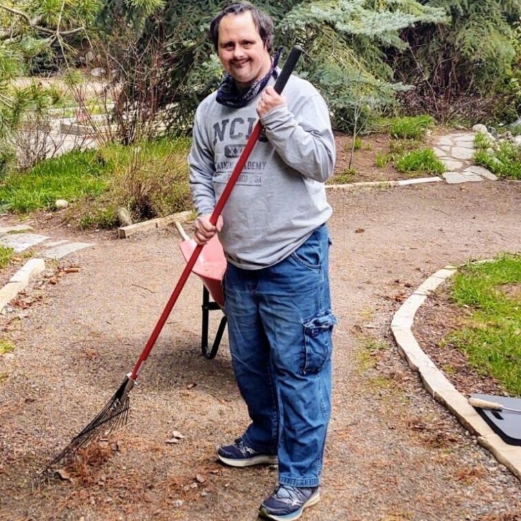 A man in a gray sweatshirt and jeans uses a rake to clean up a path in the botanic park