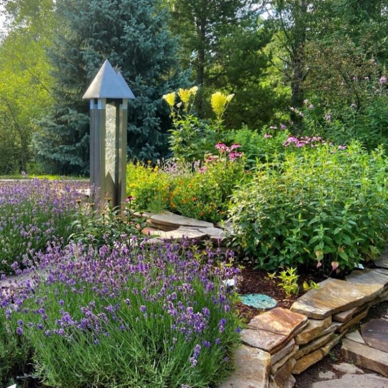 A raised garden bed containing large green herb bushes and wildflowers with a metal obelisk-like sculpture set in the middle.