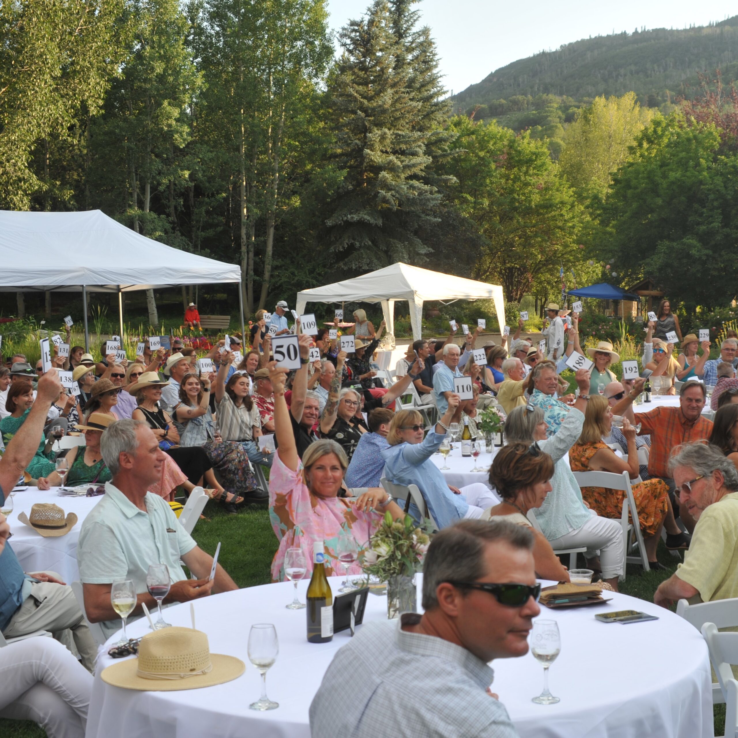 A large group of people sitting outside at tables covered with white table cloths and flower centerpieces participating in an auction