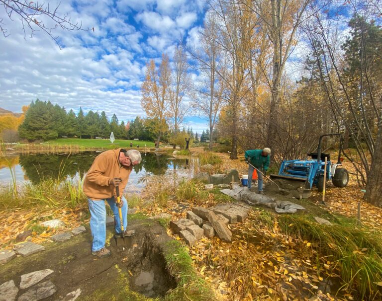 Two people digging and working with tools beside a scenic autumn pond lined with trees and fallen leaves.