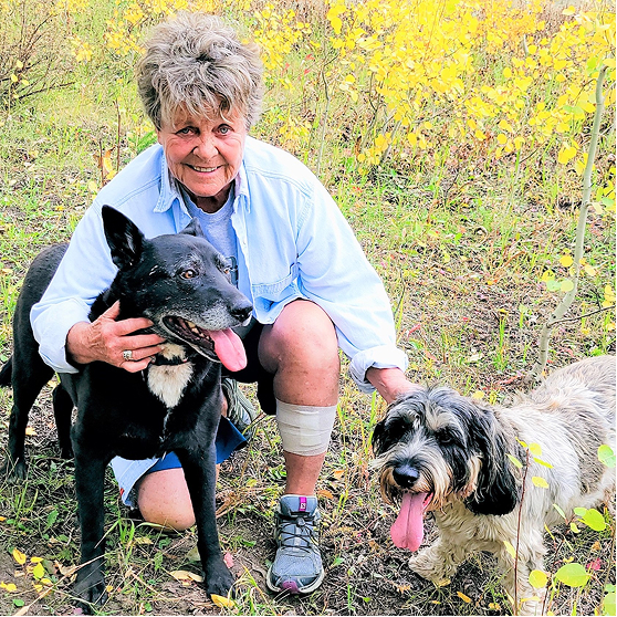 Woman in a blue shirt kneels next to two dogs with aspen trees in the background