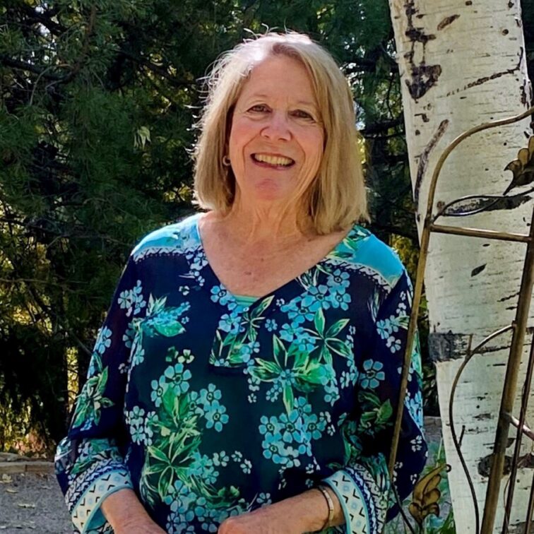 A blond woman wearing a turquoise floral shirt stands next to an aspen tree
