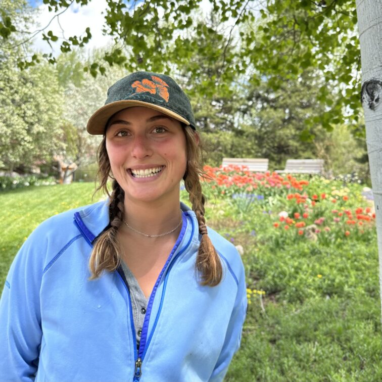 A woman in a bright blue jacket stands smiling next to a garden of wildflowers