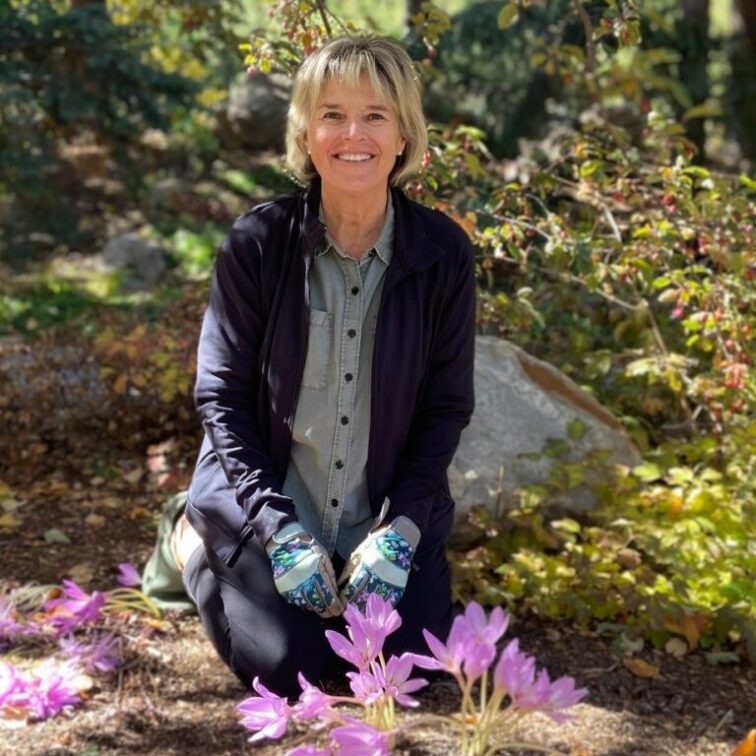A blond woman with gardening gloves on smiles as she crouches next to a cluster of pink flowers