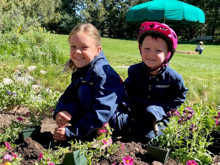 Two young children in blue jackets planting flowers in a garden bed, with one wearing a pink bike helmet.