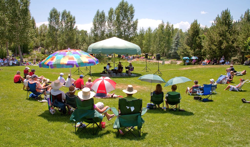 Several small groups of people underneath large colorful umbrellas sit and listen to a band play in a large grassy field on a sunny day