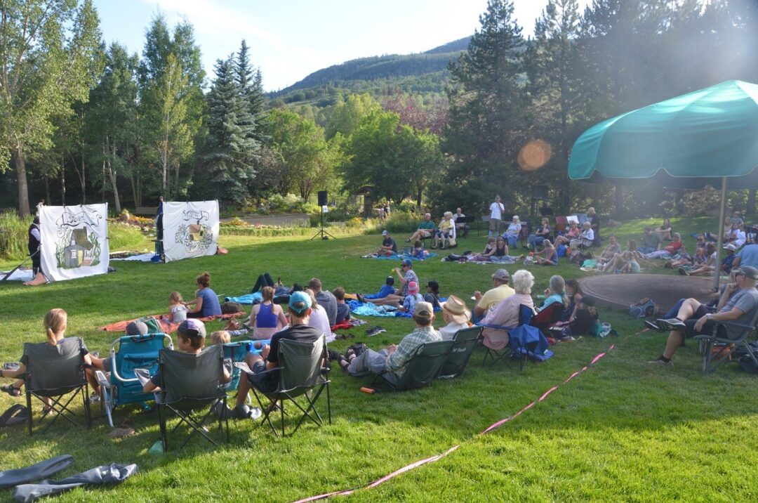 Audience members sit on blankets and lawn chairs at the Yampa River Botanic Park, watching an outdoor Piknik Theatre performance on a sunny day.