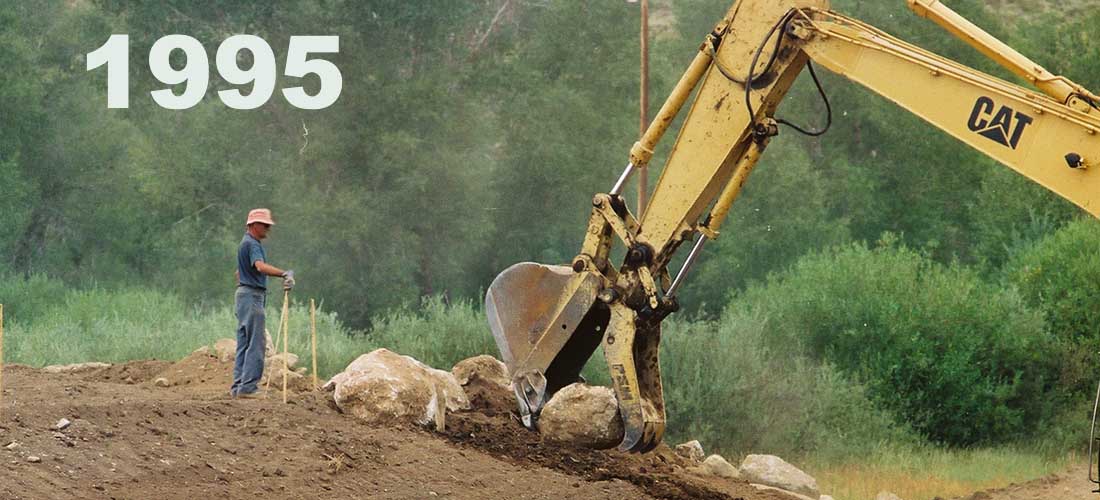 A man stands and supervises as a bulldozer picks up and moves large boulders