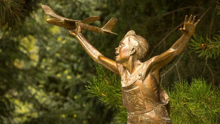 A gleaming statue that appears to be made of gold of a young boy in overalls playing with a toy plane